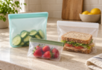 Three resealable food bags hold cucumber slices, a sandwich and strawberries on a kitchen counter.