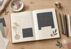 An open scrapbook with blank cream and charcoal paper, four unbranded pens, paper cutouts and decorative tape on a wooden table