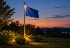 A blue flag on a silver pole with a small light beneath the finial at dusk.