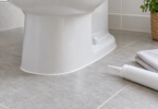 A white toilet base with a narrow white bead along a gray tile floor, beside an unbranded sealant container and a folded cloth