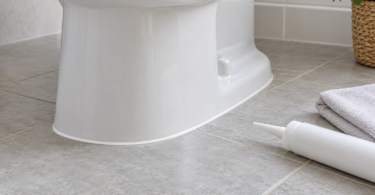 A white toilet base with a narrow white bead along a gray tile floor, beside an unbranded sealant container and a folded cloth