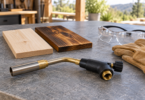 An unlit, disconnected torch head lies on a gray work surface beside pale and dark-grained wood samples, safety glasses and leather gloves.