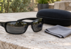 Black wraparound sunglasses beside a closed black case and folded gray cleaning cloth on a stone table.