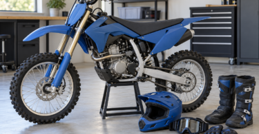 Unbranded blue dirt bike on a stand beside a full-face helmet, goggles, gloves and tall riding boots in a garage.